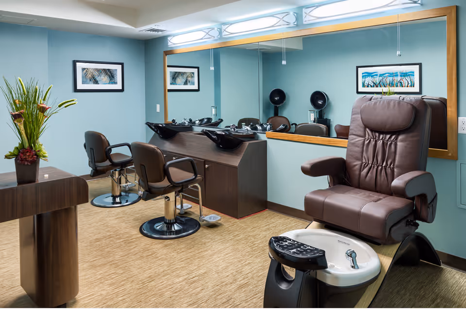 Interior of a salon area in Kalākaua Gardens featuring two brown salon chairs in front of wash basins, a large mirror on the wall, a comfortable brown pedicure chair with a foot bath, and a table with a flower arrangement. The walls are painted light blue and decorated with framed artwork.