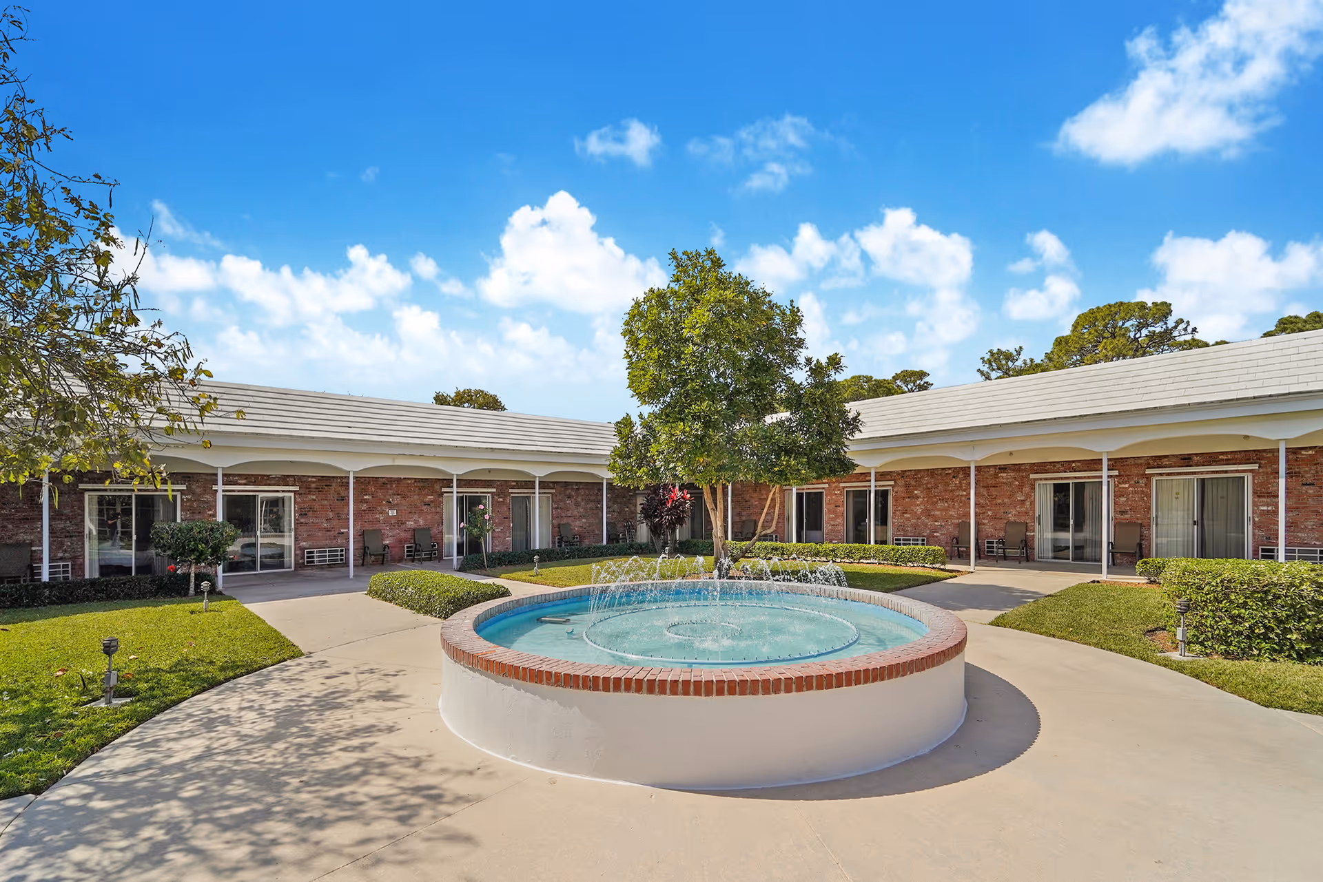 Outdoor courtyard area of Manor Pines facility featuring a circular water fountain with water jets, surrounded by a paved walkway, green grass, bushes, and trees. The courtyard is enclosed by a single-story brick building with multiple sliding glass doors and covered walkways. The sky is blue with scattered white clouds.