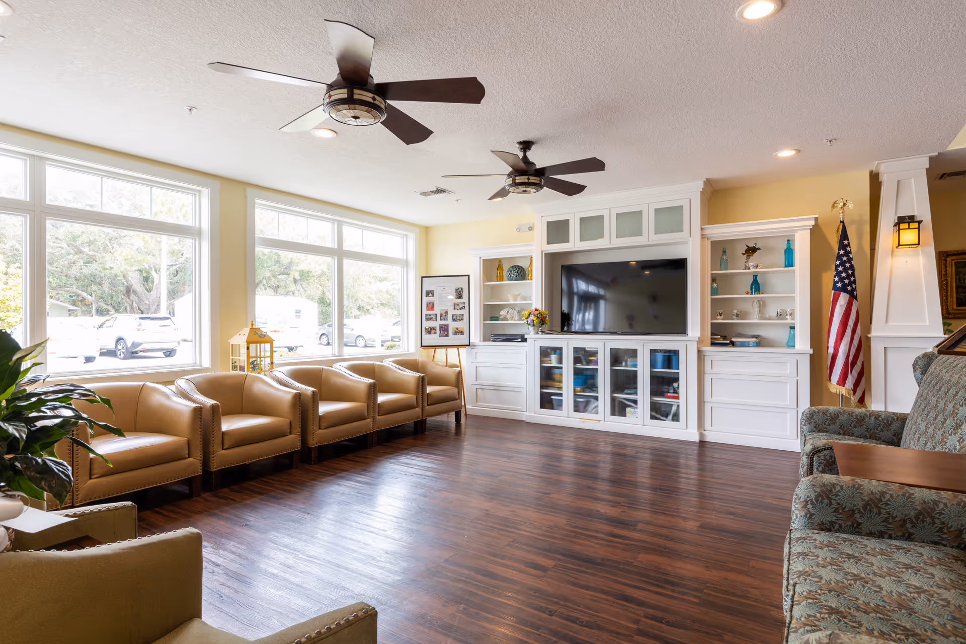 A spacious living room with large windows letting in natural light. The room features a row of tan leather armchairs along the window wall, two ceiling fans, a large flat-screen TV mounted on a white built-in cabinet with shelves, and an American flag in the corner. The floor is dark wood, and there are additional upholstered chairs with floral patterns on the right side.