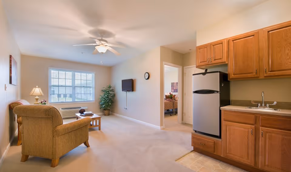 A cozy living area in a senior living facility with a beige armchair, a wooden coffee table, a lamp on a side table, a wall-mounted TV, a ceiling fan, a large window with blinds, and a small kitchenette with wooden cabinets, a sink, and a stainless steel refrigerator. A doorway leads to a bedroom in the background.