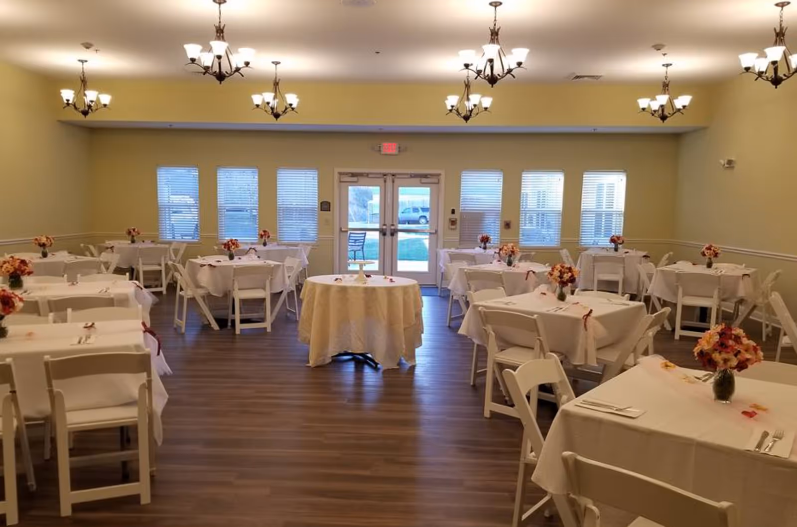 A dining room with multiple tables covered in white tablecloths, each set with cutlery and decorated with small flower arrangements. The room has wooden flooring, yellow walls, several windows with blinds, and multiple chandeliers hanging from the ceiling. Double glass doors are visible at the far end of the room.