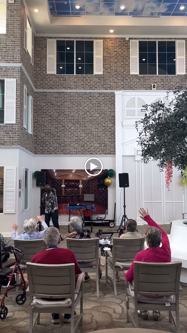 A group of elderly people seated in chairs facing a person standing in front of a backdrop that looks like a wine cellar. The setting is indoors with a high ceiling, brick-patterned walls, large windows, and a decorative tree on the right. Some of the seated individuals have their hands raised.