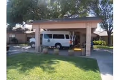 Covered carport/entrance of a single-story facility with a white van parked underneath, a grassy lawn and trees in front.