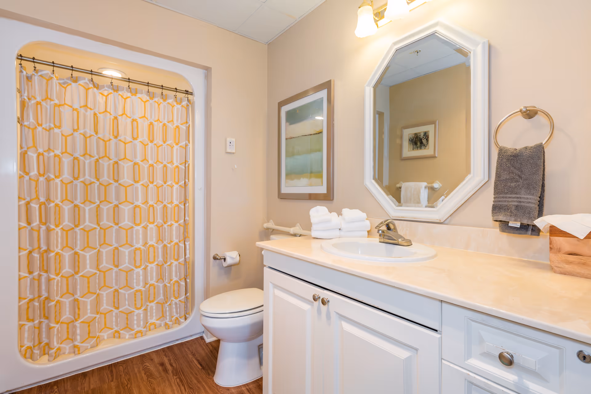 A clean bathroom with a white toilet, a sink with a beige countertop, a white cabinet below, and a mirror above the sink. There is a yellow and white patterned shower curtain on the left side, a towel ring with a gray towel on the right wall, a framed abstract painting on the wall, and a wooden floor.