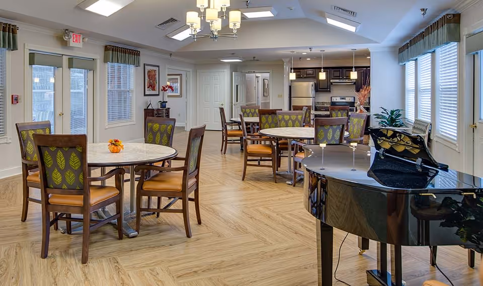 A spacious and well-lit common area with multiple round tables and chairs arranged for seating. The room features large windows with blinds and valances, a black grand piano on the right side, and a kitchen area with stainless steel appliances in the background. The floor has a light wood pattern, and there are ceiling lights and a chandelier providing illumination.