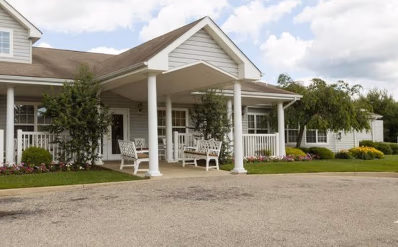 Exterior view of a single-story assisted living facility with a covered entrance supported by white columns. There are white benches on the porch, surrounded by green shrubs and colorful flowers. The sky is partly cloudy.