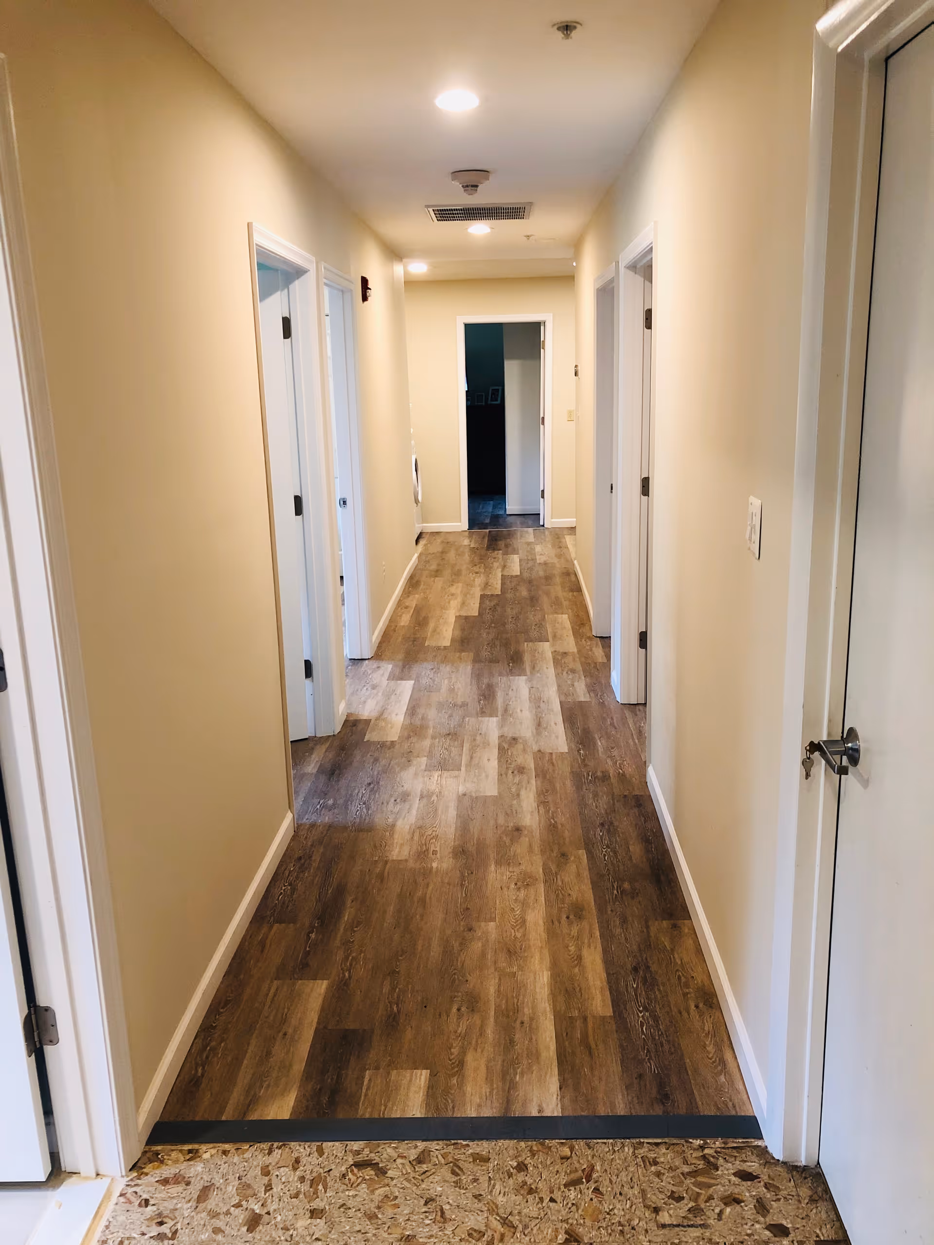 A long, narrow hallway with light beige walls and wood-patterned flooring. There are multiple white doors on both sides of the hallway, and recessed ceiling lights illuminate the space.