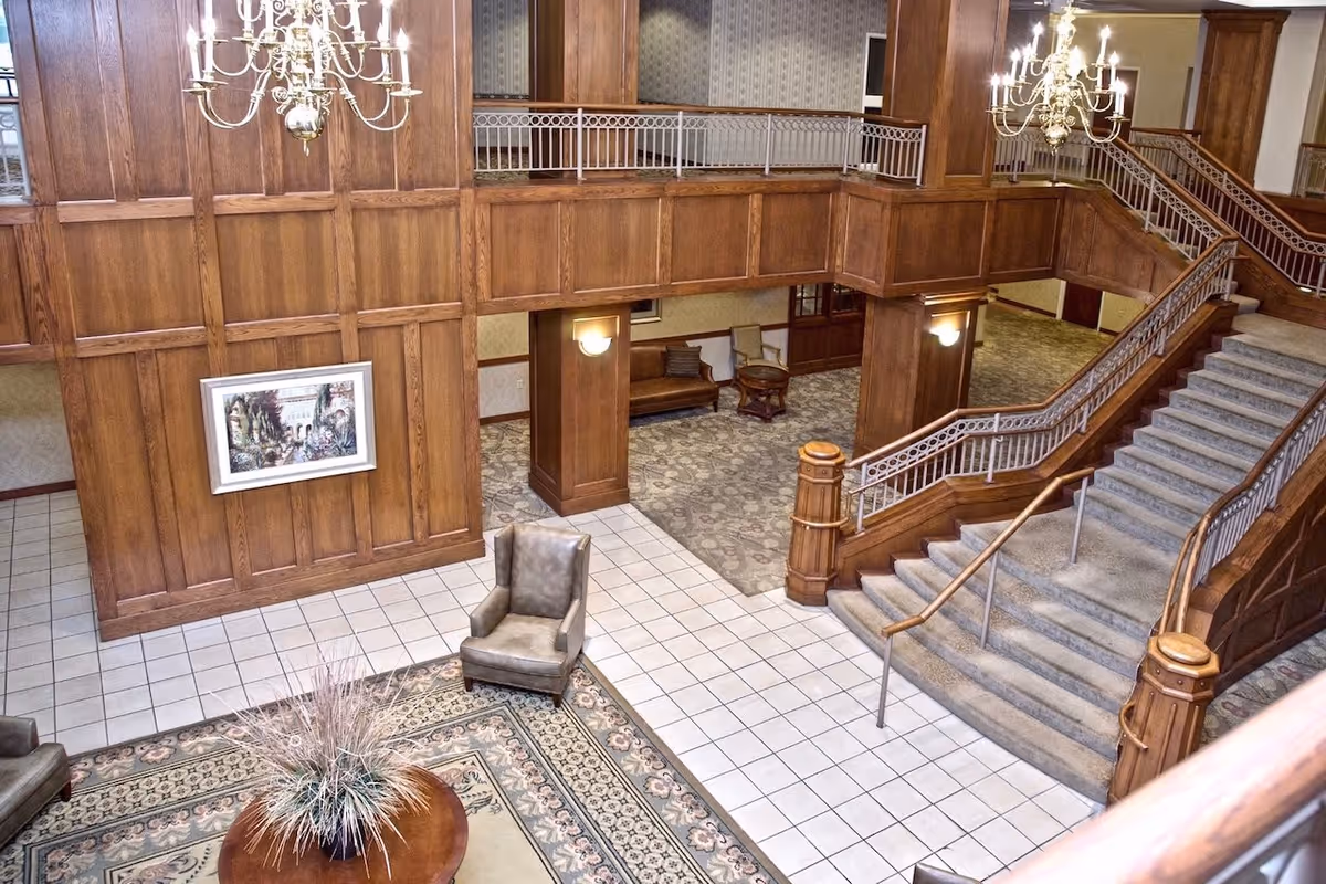 Interior view of a senior living facility lobby with a large wooden staircase, two chandeliers, wood-paneled walls, a framed painting, armchairs, a round table with a plant, and carpeted and tiled flooring.