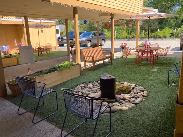 Outdoor seating area with artificial grass, a circular stone fire pit, metal chairs, wooden bench, and tables with umbrellas. The area is covered by a roof and surrounded by planters and parked cars in the background.