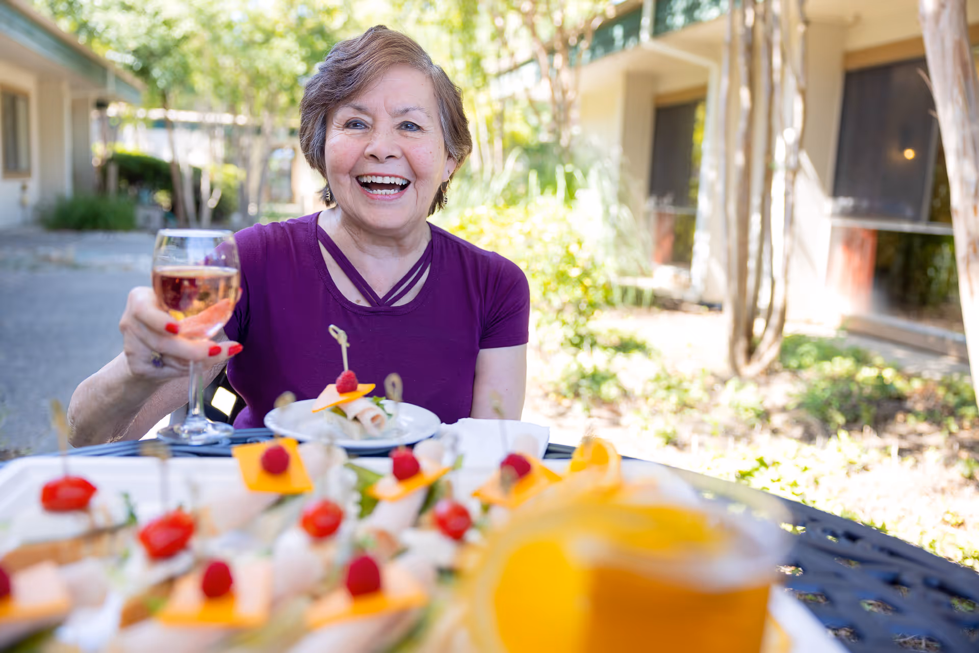A smiling elderly woman wearing a purple shirt is sitting outdoors at a table with appetizers and holding up a glass of rosé wine. The background shows greenery and the exterior of a building.