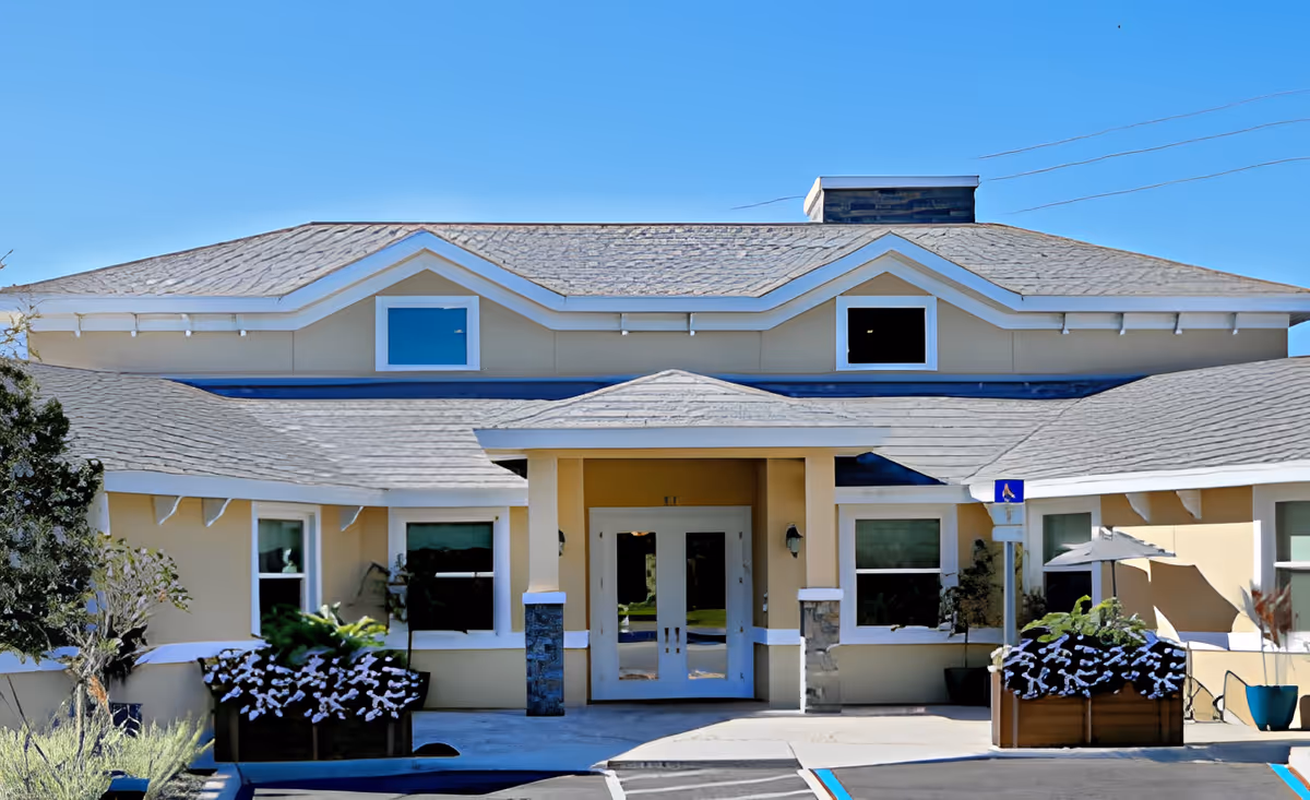 Front exterior view of a single-story building with beige walls and a gray shingled roof under a clear blue sky. The entrance has double glass doors with a small covered porch supported by two columns. There are windows on either side of the entrance and flower planters with white flowers near the walkway. A handicapped parking sign is visible near the entrance.