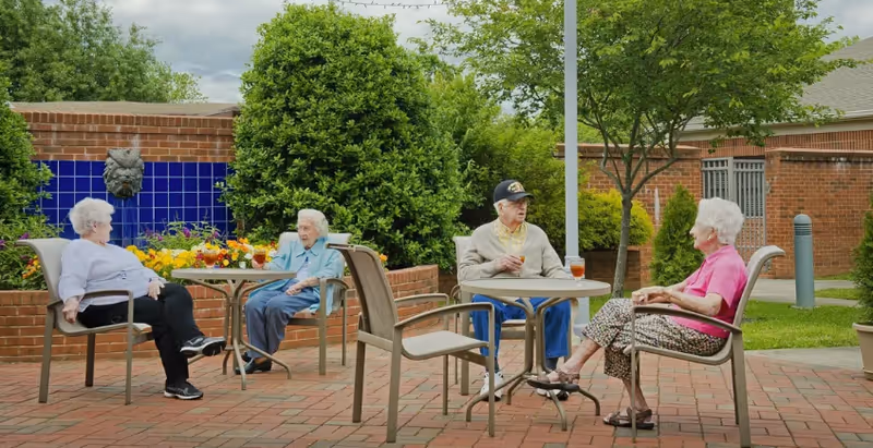 Four elderly residents sitting at patio tables and talking in a brick-paved courtyard with plants and a tiled water feature.