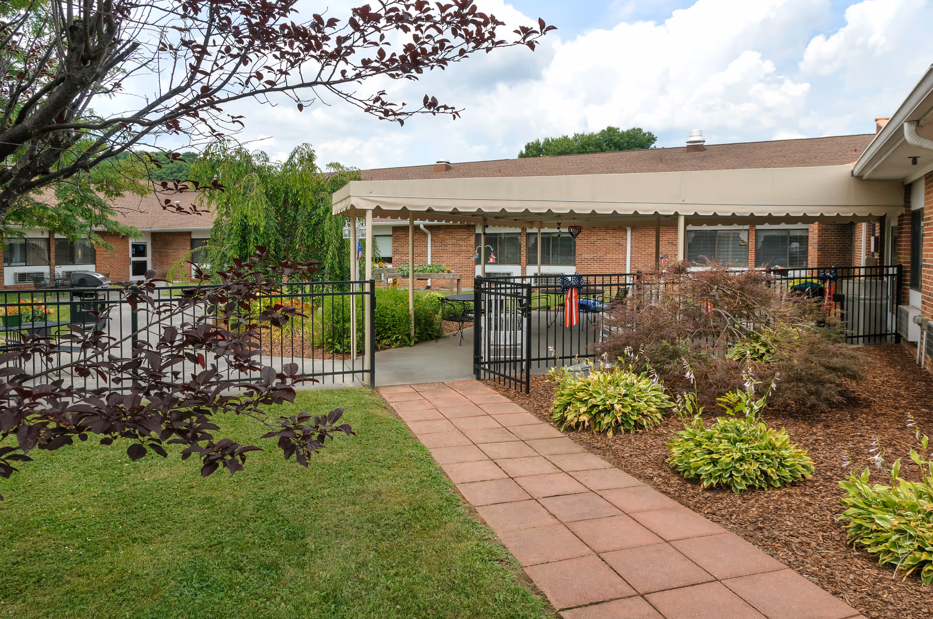 A landscaped outdoor courtyard with a paved walkway leading to a fenced patio and single-story brick building under a cloudy sky.