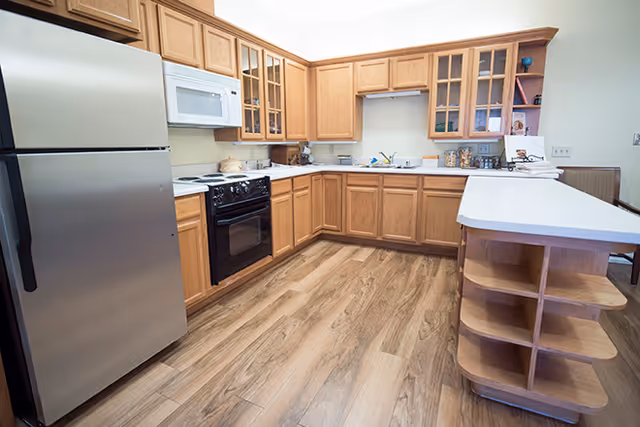 Kitchen with light wood cabinets, stainless steel refrigerator, stove and microwave, and a white countertop peninsula with open shelving.