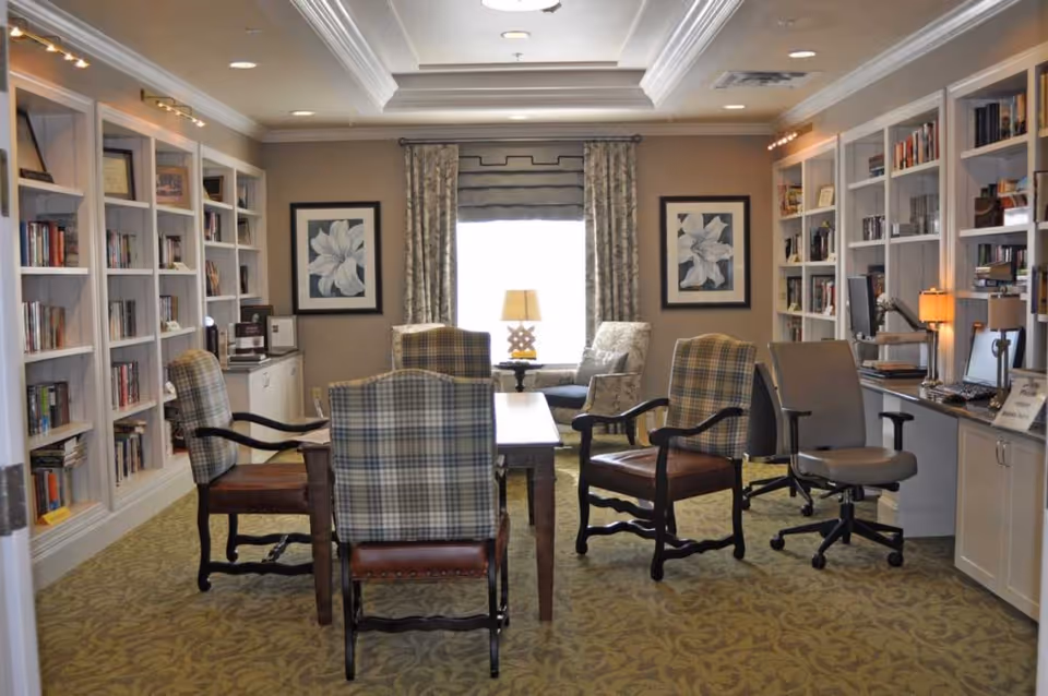 A cozy common room/library with bookshelves lining the walls, a central table surrounded by upholstered chairs, lamps and computer workstations.