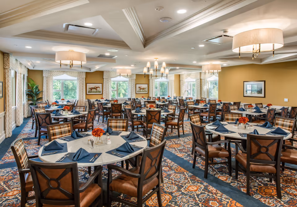 A spacious dining room with multiple round and rectangular tables set with blue napkins, silverware, and small floral centerpieces. The room features patterned carpet, large windows with curtains, framed artwork on the walls, and several ceiling light fixtures including chandeliers and lamps.