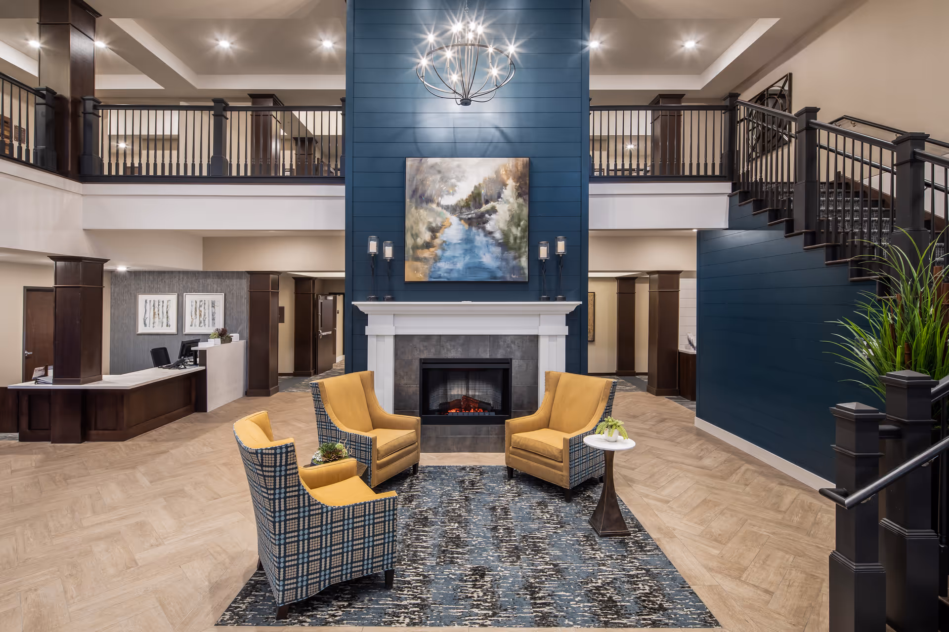A spacious and elegant senior living facility lobby with a high ceiling and a modern chandelier. The focal point is a blue accent wall with a fireplace and a landscape painting above it. In front of the fireplace are four yellow and plaid armchairs arranged around a blue patterned rug with a small side table holding a plant. To the left is a reception desk with two chairs and decorative artwork on the wall behind it. A staircase with dark wood railings leads to an upper balcony overlooking the lobby. The floor is light wood, and the overall decor is warm and inviting.