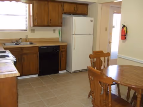A kitchen area with wooden cabinets, a white refrigerator, a black dishwasher, a sink under a window, and a wooden dining table with chairs. A fire extinguisher is mounted on the wall near a door with a window.