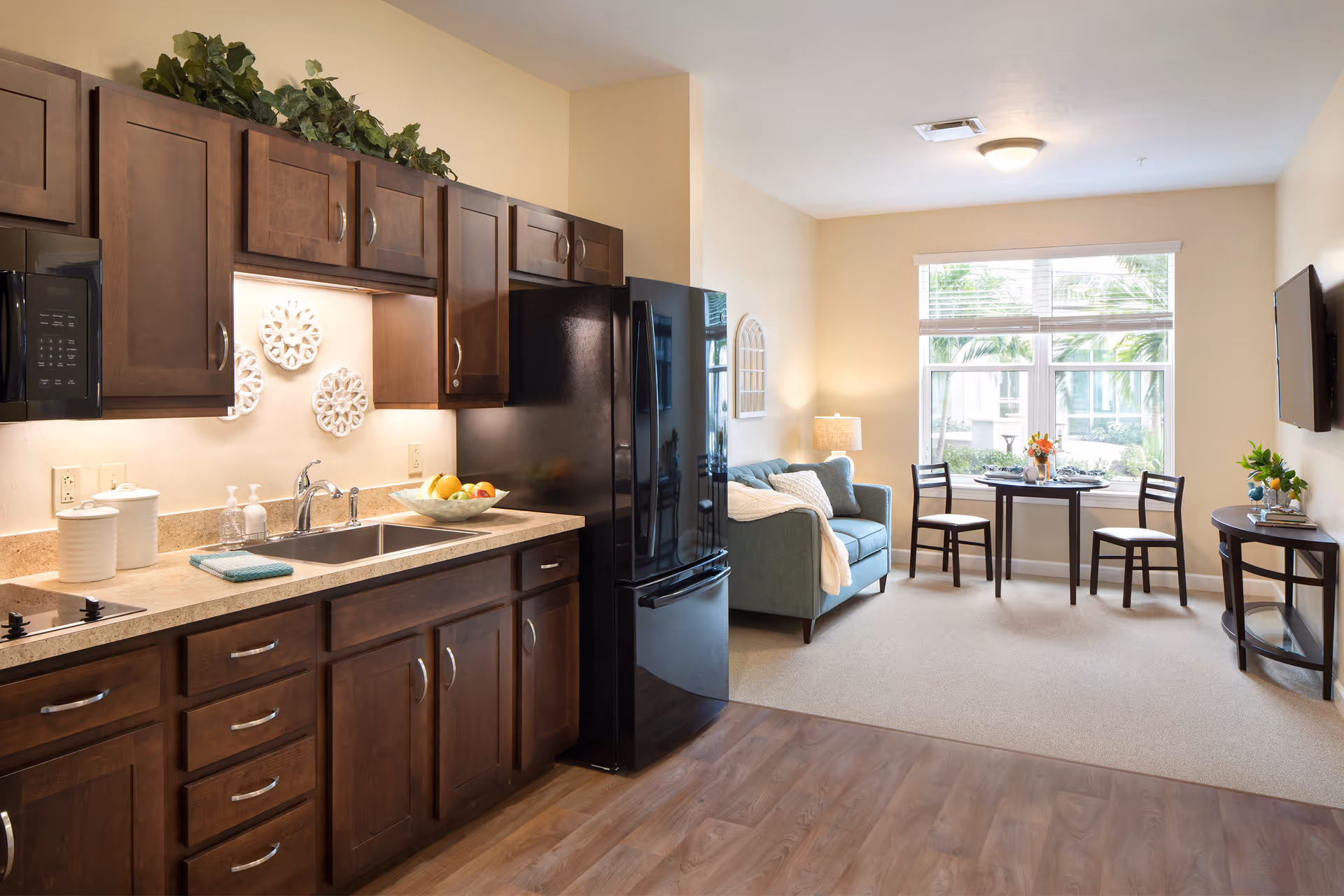 A bright and cozy living space featuring a kitchen area with dark wood cabinets, a black refrigerator, and a sink with a bowl of fruit on the counter. Adjacent to the kitchen is a small living room with a blue loveseat, a round dining table with two chairs near a large window, and a wall-mounted TV. The room has light-colored walls and carpeted flooring in the living area, with wood flooring in the kitchen.
