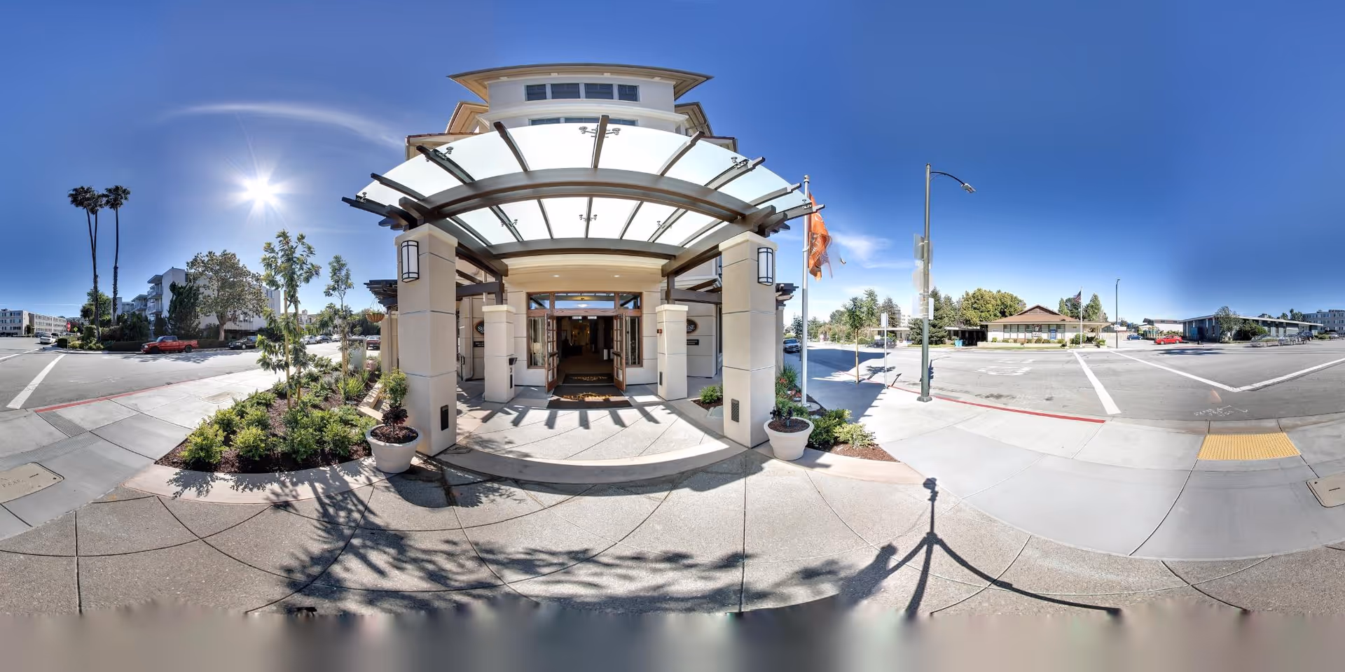 Wide-angle view of the main entrance to Oakmont of Burlingame senior living facility on a sunny day, showing a covered entryway with glass canopy, potted plants, and surrounding sidewalks and streets with clear blue sky.