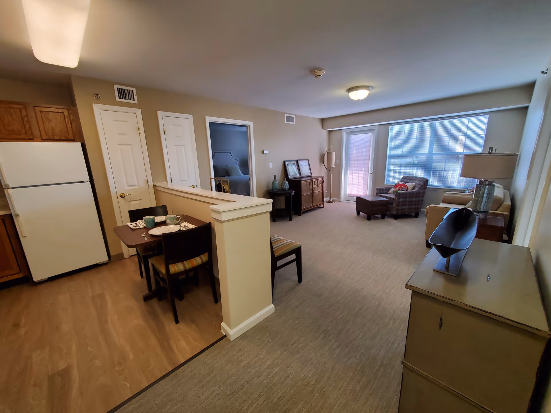 Interior view of a senior living facility apartment showing a small dining area with a table set for two, a kitchen with a refrigerator and wooden cabinets, and a living room with a chair, ottoman, sofa, and large window with blinds. There are two closed doors and an open doorway leading to a bedroom.