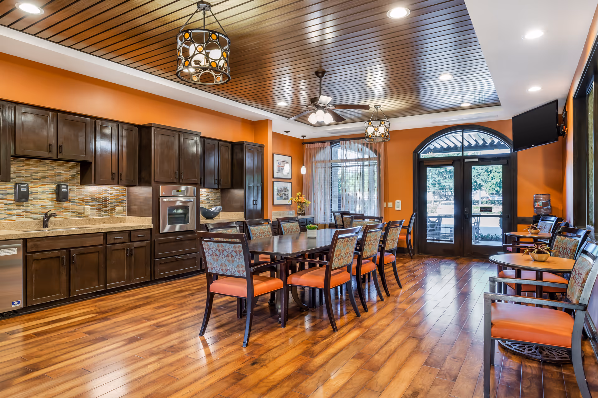 Bright communal dining area with a long wooden table and chairs, adjacent kitchen cabinets and double glass doors to the outside.