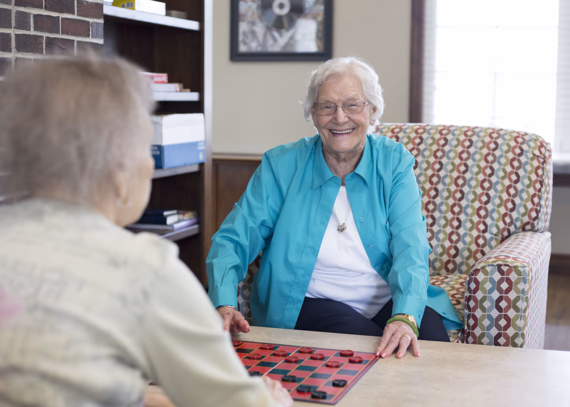 Two elderly women sitting at a table playing checkers in a cozy room. One woman with white hair and glasses is smiling and wearing a turquoise shirt, while the other woman is seen from behind. The room has a bookshelf and a window with blinds.
