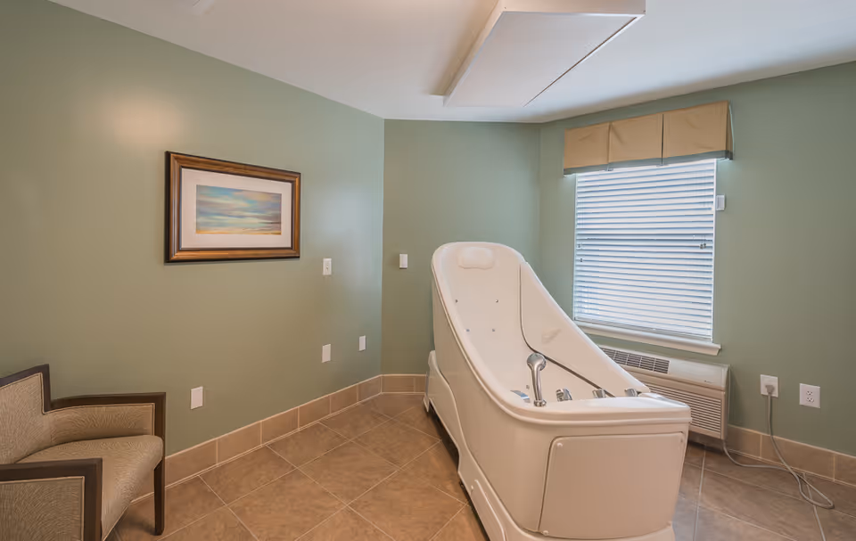 Small tiled room with a medical-style white bathing tub, a chair, framed artwork on a green wall, and a window with blinds.