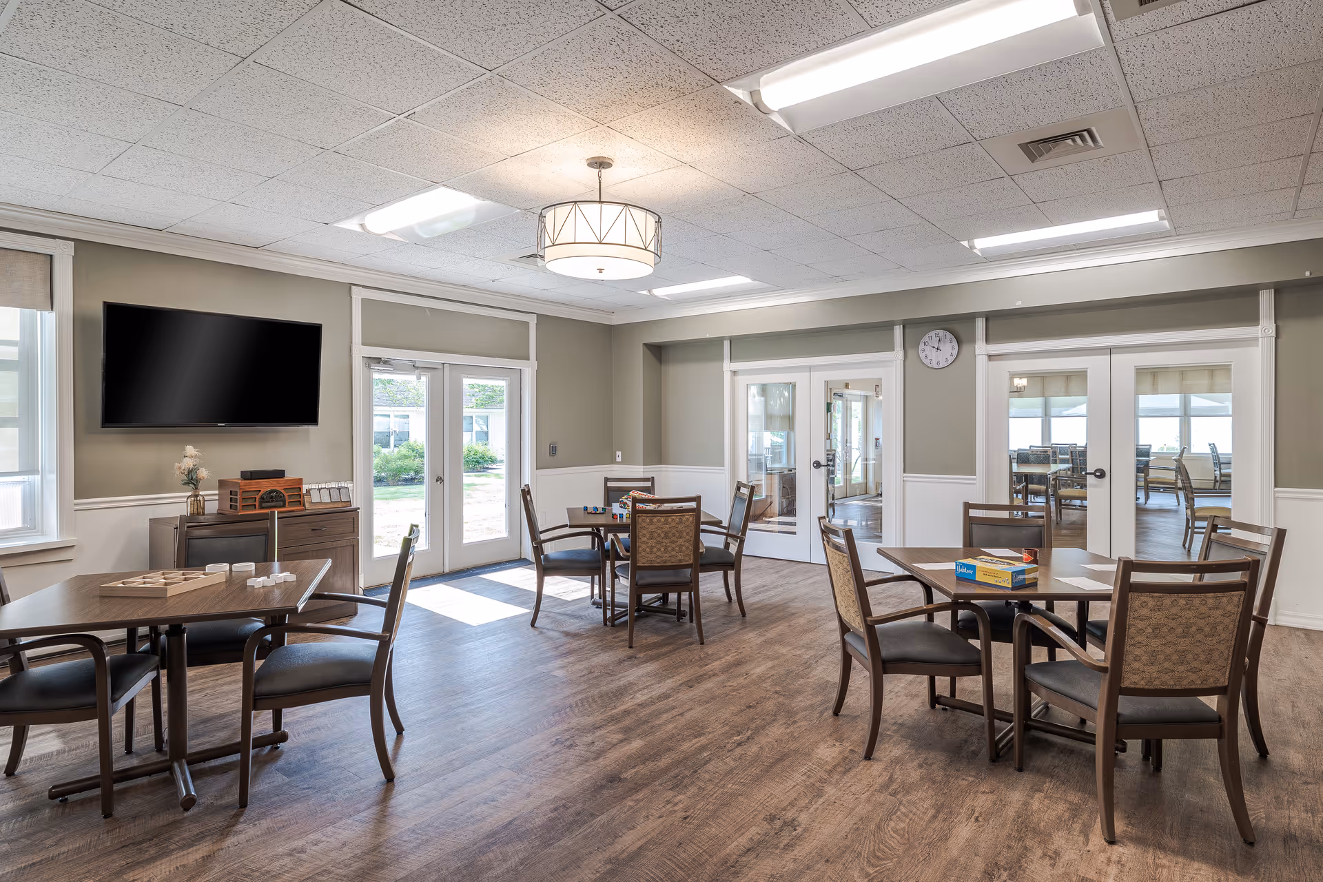 Bright common activity/dining room with multiple tables and chairs, a wall-mounted TV, and glass doors to an outdoor area.