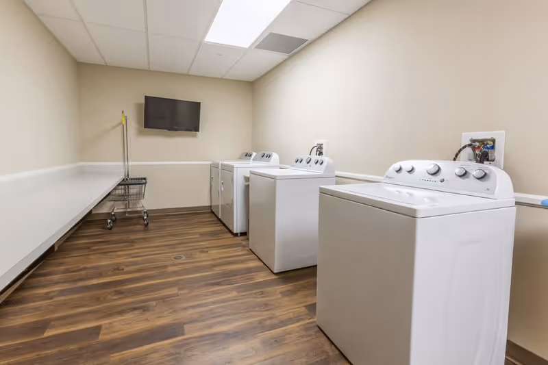Laundry room with multiple washing machines and dryers, a long folding counter, a utility cart, and a wall-mounted TV.