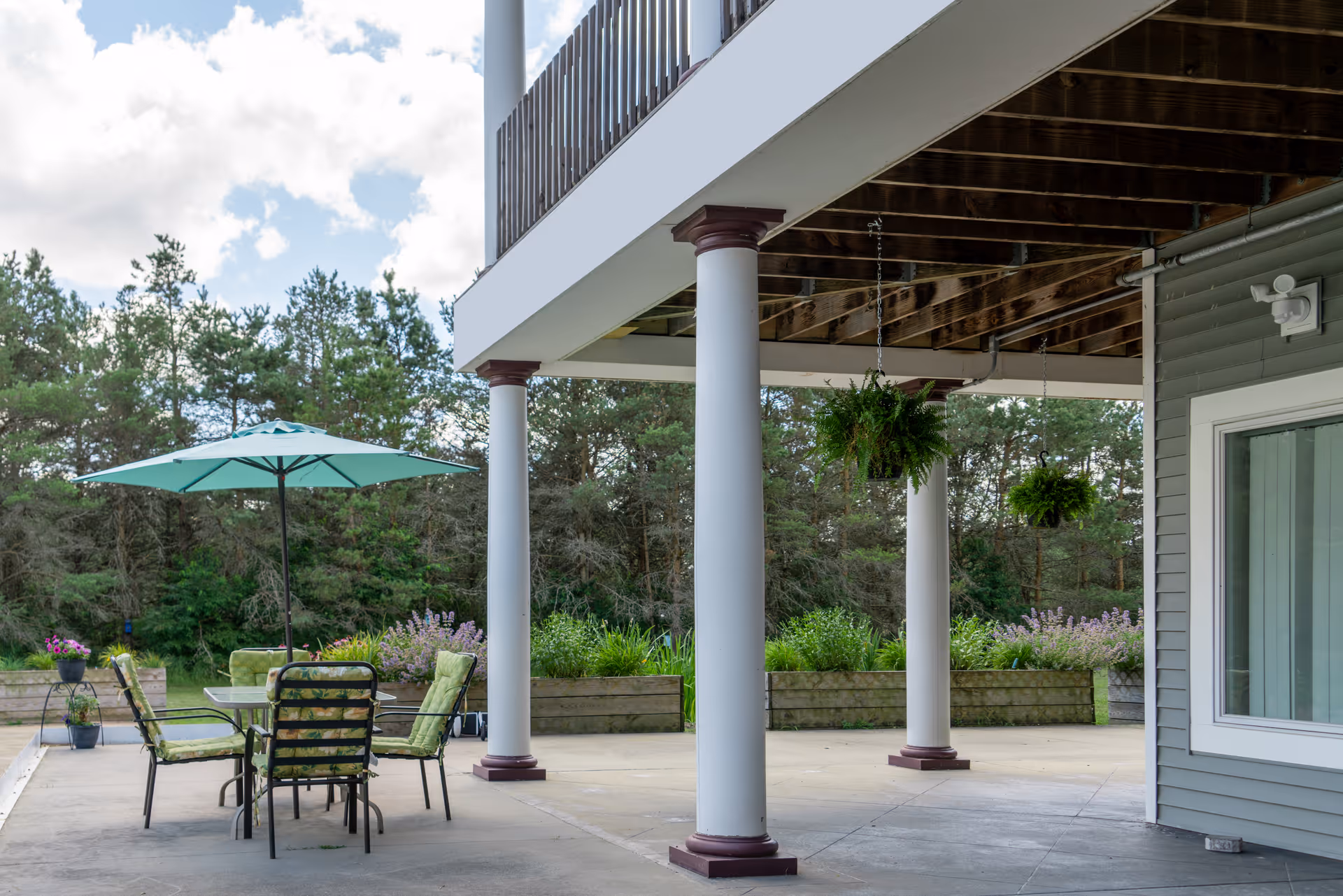 Outdoor patio area at Whispering Pines Senior Living with a glass table, four cushioned chairs, and a large teal umbrella. The patio is supported by white columns with brown bases and tops, hanging green plants, and surrounded by greenery and trees under a partly cloudy sky.