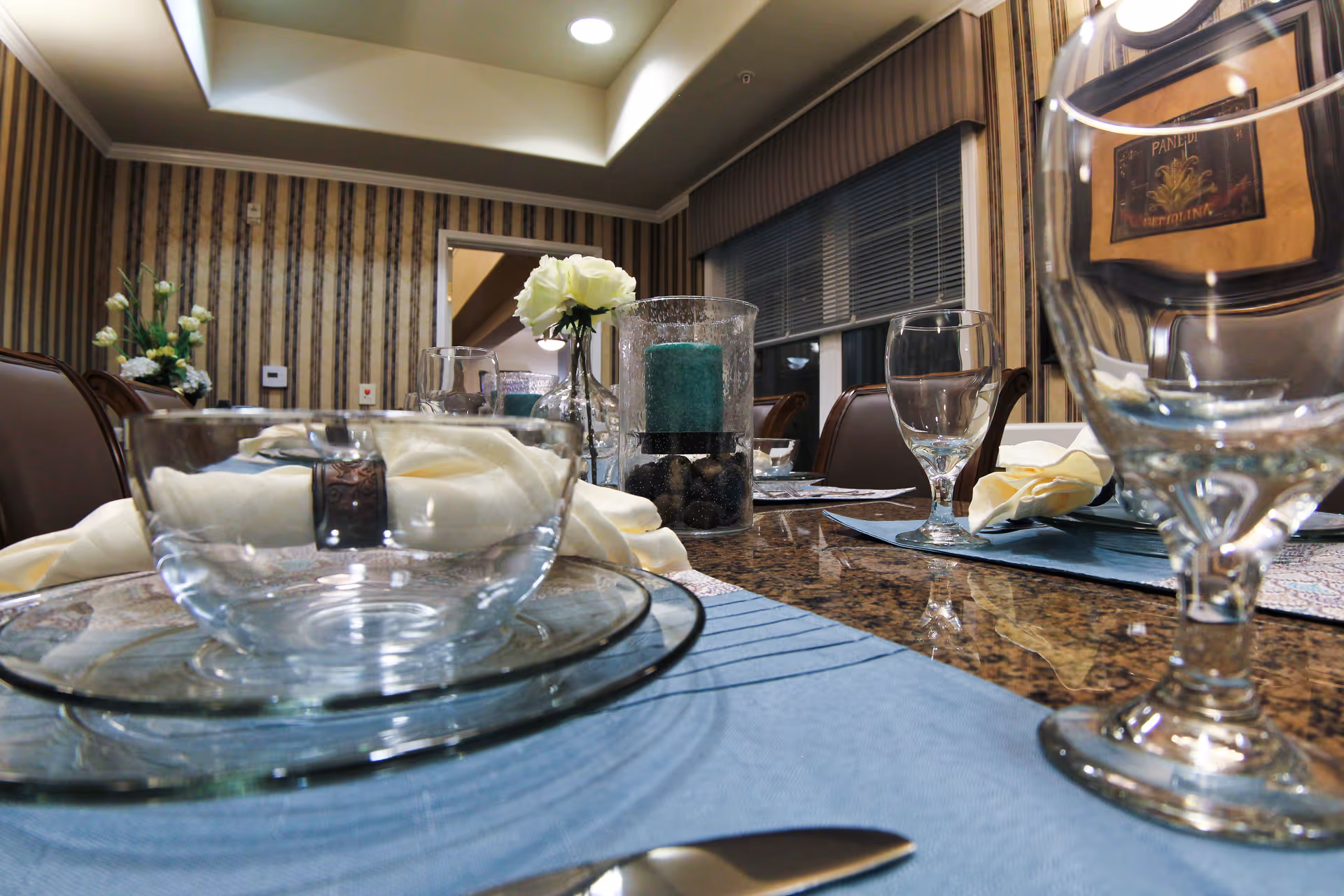 Close-up view of a dining table set with clear glass bowls, plates, water glasses, and cream-colored napkins on blue placemats in a room with striped wallpaper and a window with blinds. A centerpiece with a blue candle and white flowers is also visible.