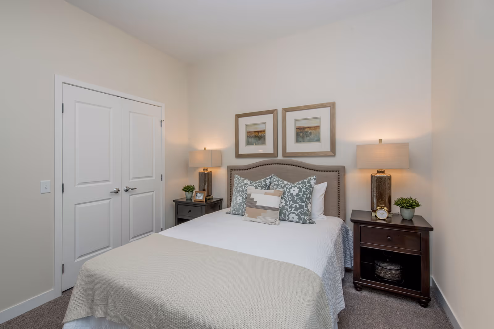 A neatly made bed with a beige headboard, white bedding, and decorative pillows in a bedroom. There are two dark wooden nightstands on either side of the bed, each with a lamp, a small plant, and decorative items. Two framed artworks hang on the wall above the bed. A closed white double-door closet is visible to the left.