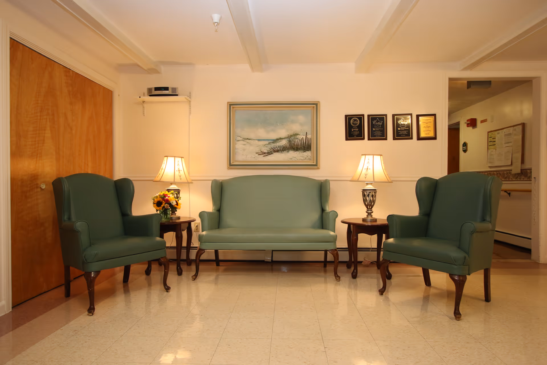 A sitting area with two green upholstered armchairs and a matching green upholstered loveseat arranged around two wooden side tables with lamps. A framed painting of a beach scene and four plaques are mounted on the wall behind the seating. The floor is tiled, and there is a wooden door on the left side of the image.