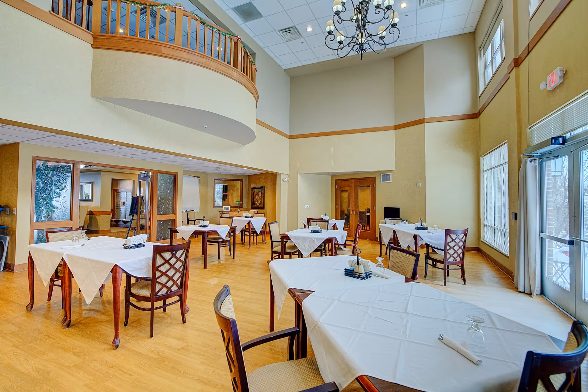 A spacious dining room with several tables covered in white tablecloths, each set with napkins and glasses. The room features high ceilings with a chandelier, large windows letting in natural light, wooden floors, and a balcony railing above. There are double doors and an exit door visible in the background.