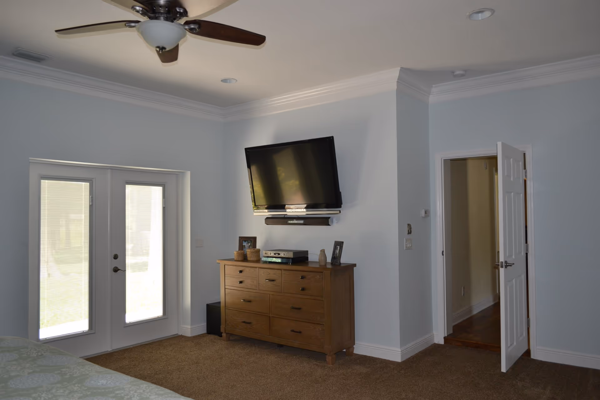 Bright bedroom interior with a wall-mounted TV above a wooden dresser, ceiling fan, and French doors leading outside.