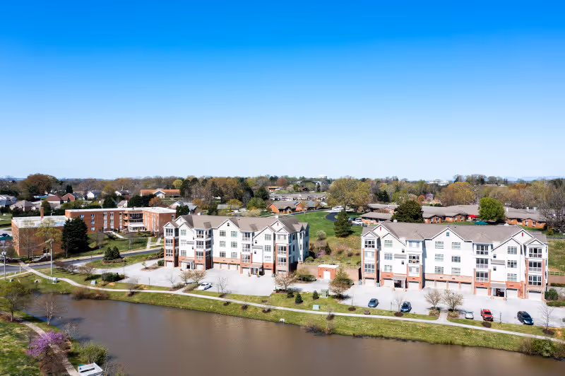 Aerial view of Asbury Place Maryville senior living facility showing multiple multi-story residential buildings near a body of water with parking lots and green spaces around. Trees and other residential houses are visible in the background under a clear blue sky.