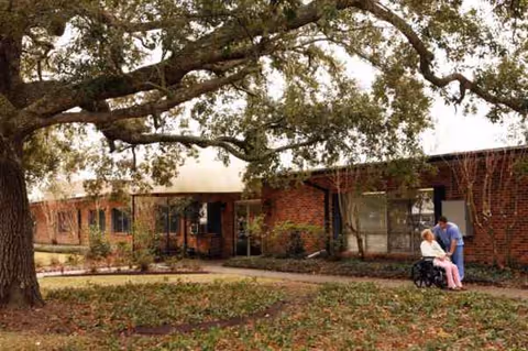 Exterior view of New Iberia Manor South showing a tree, walkway, and caregiver with resident.