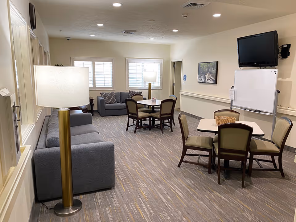 A well-lit common area in an assisted living facility featuring two gray sofas, two tables each surrounded by four chairs, a floor lamp, a wall-mounted TV, and a whiteboard. The room has carpeted flooring, beige walls, and windows with white shutters allowing natural light in.