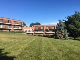 A large grassy lawn with a few trees in front of a multi-story residential building under a clear blue sky.