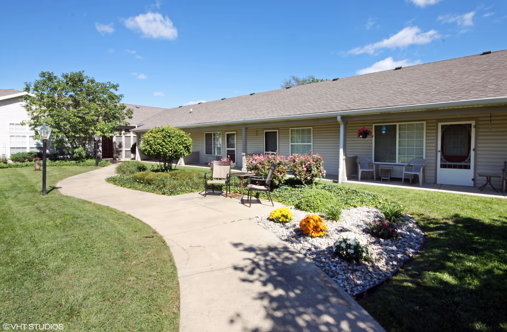 Exterior courtyard of a single-story Riverbend senior living building with a curved sidewalk, lawn, patio chairs and flower beds.