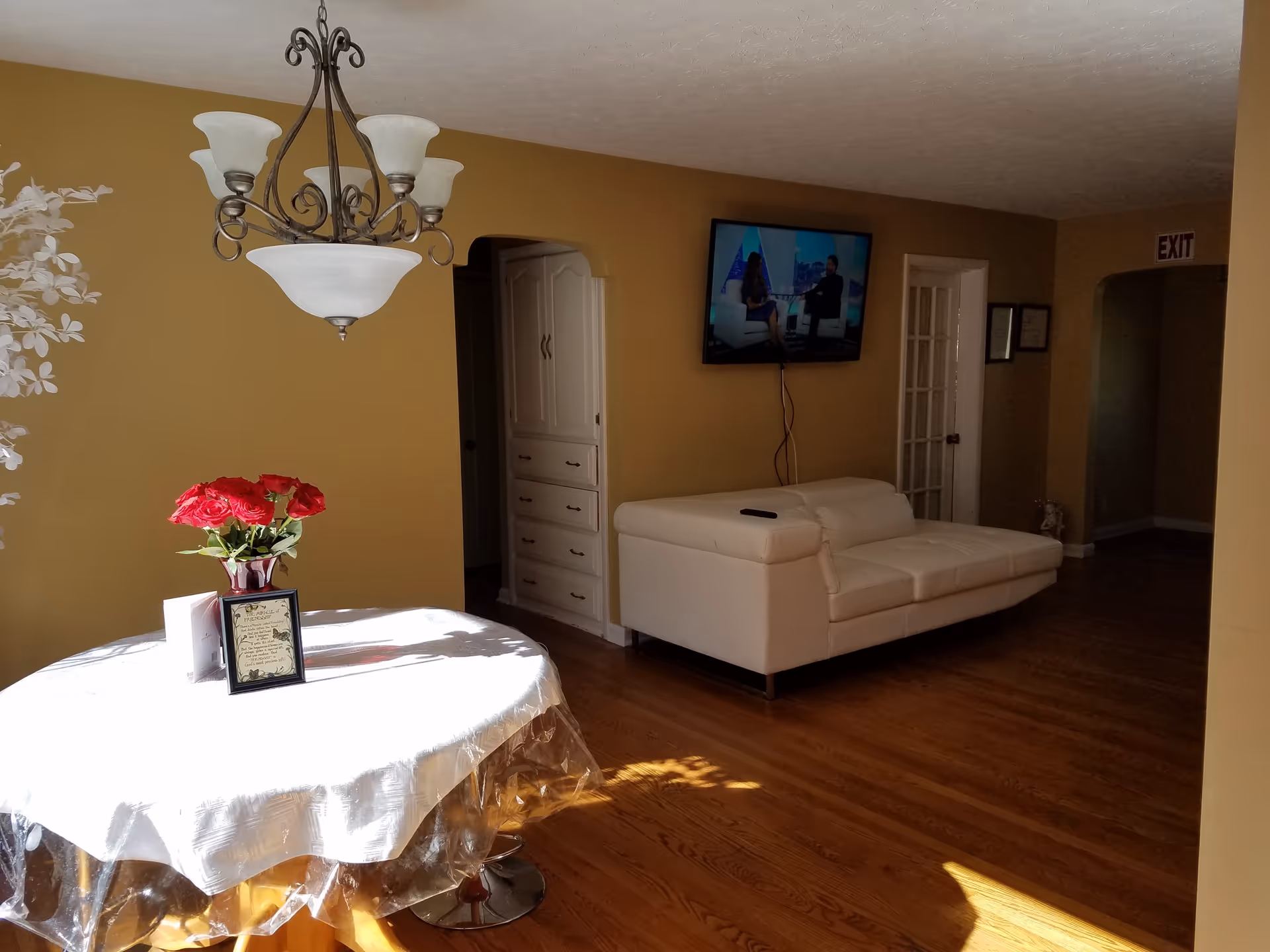 Bright interior room with a round table topped with red flowers under a chandelier and a white sofa facing a wall-mounted TV.