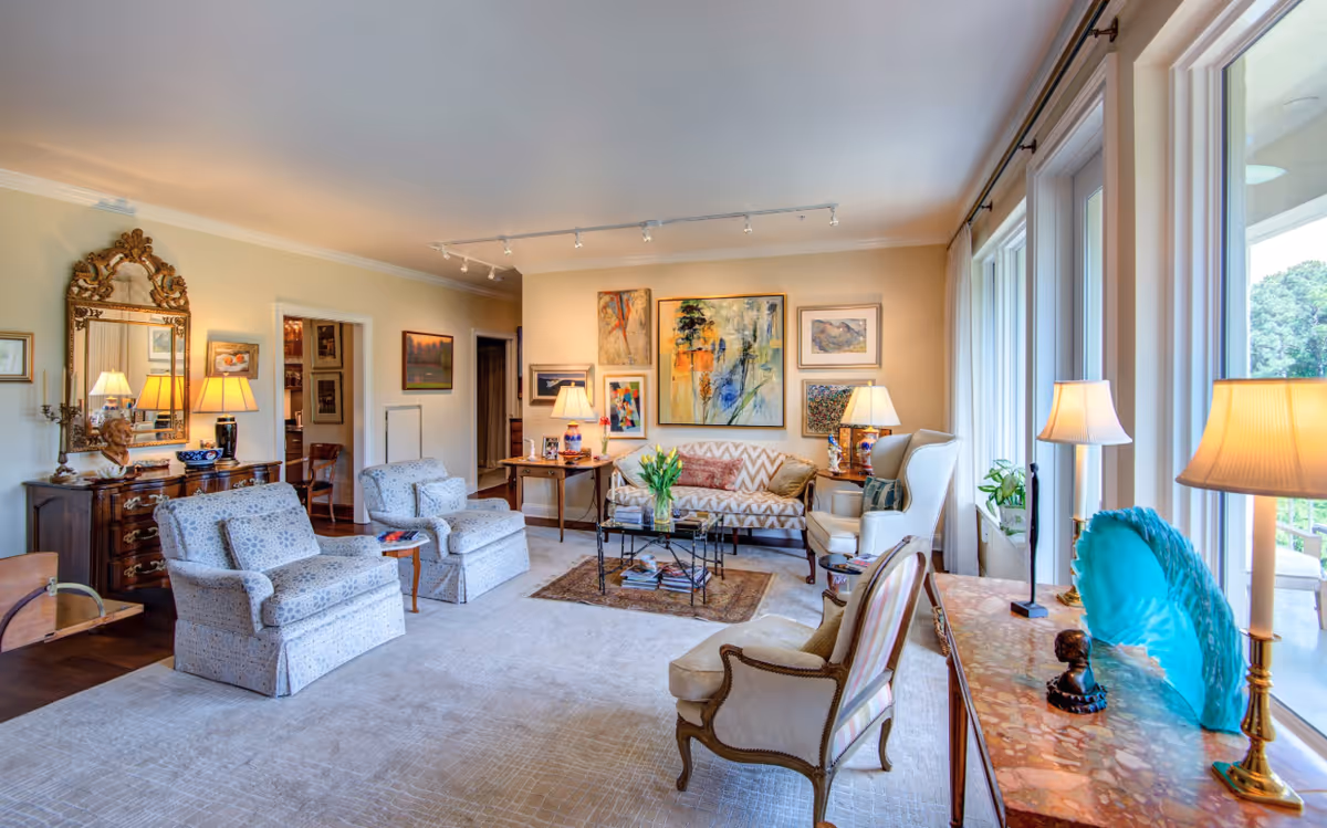 A spacious and well-lit living room with large windows on the right side letting in natural light. The room features a patterned sofa, two upholstered armchairs, a wooden side table with a lamp, and a coffee table with a vase of yellow tulips. The walls are adorned with various framed artworks. There is a decorative mirror above a wooden dresser on the left side, and several table lamps provide additional lighting. The room has a cozy and elegant ambiance with a mix of classic and contemporary decor.