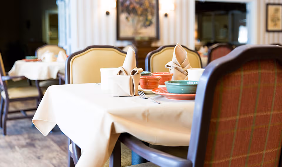 A dining room table set with beige tablecloth, folded beige napkins, colorful bowls and plates, and silverware. The chairs around the table have cushioned backs with a plaid pattern. The background shows more tables and chairs in a softly lit room with framed artwork on the walls.