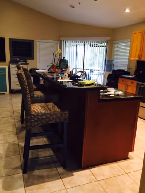 Interior view of a kitchen area with a large island countertop featuring three wicker bar stools. The kitchen has wooden cabinets, a water dispenser, and various items on the countertop. Sliding glass doors with vertical blinds lead to an outdoor area.
