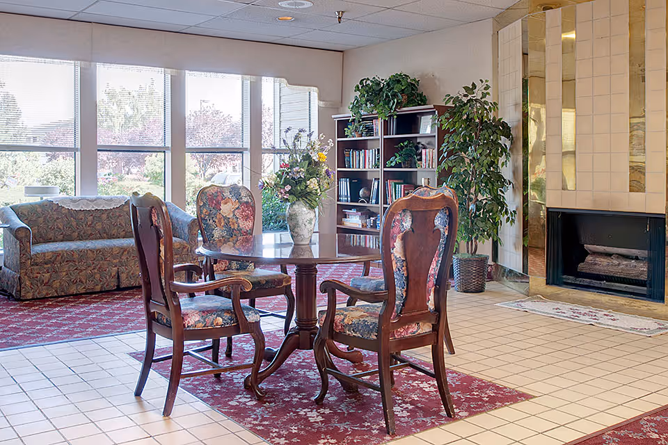Bright common room with a round wooden table surrounded by floral-upholstered chairs, a couch, bookshelf, plants, and a fireplace by large windows.