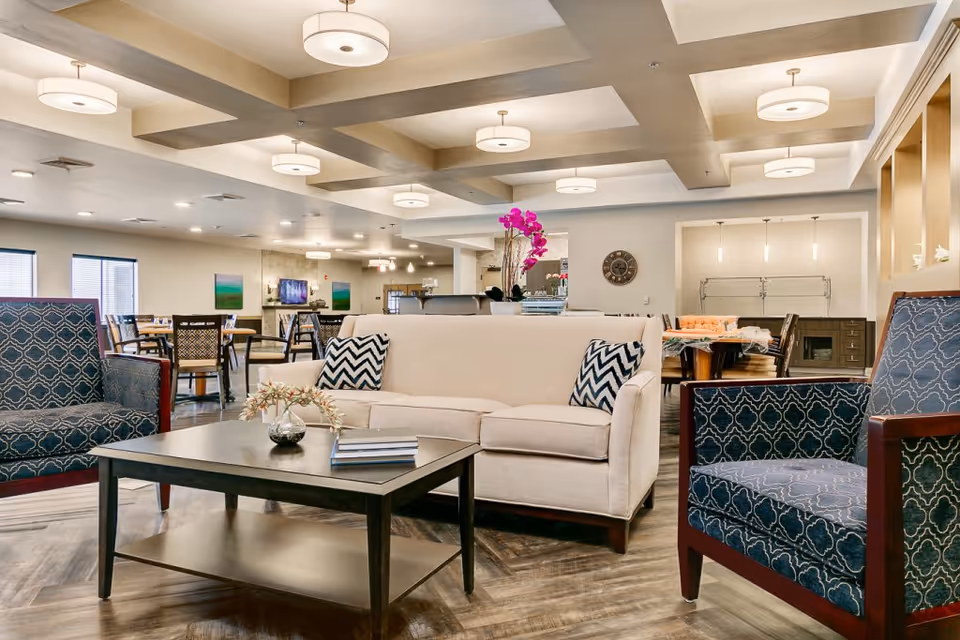 A spacious and well-lit senior living common area featuring a beige sofa with two patterned pillows, two blue patterned armchairs, and a dark wooden coffee table with a small plant and books. In the background, there are dining tables and chairs, modern ceiling lights, and decorative elements including a wall clock and a vase with pink flowers.
