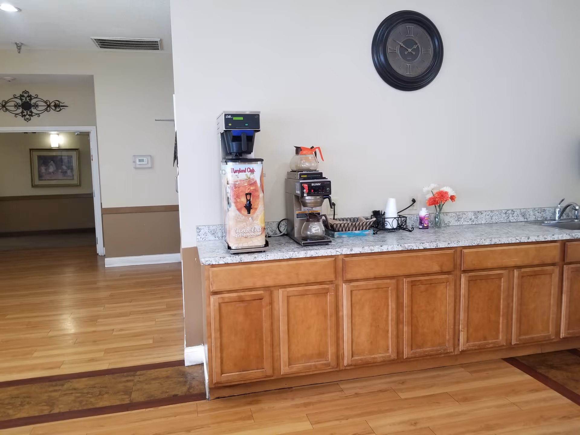 A countertop area with a coffee maker, iced tea dispenser, and a small sink. There are wooden cabinets below the countertop and a round wall clock above. The floor is wood, and there is an open doorway leading to another room with a framed picture on the wall.