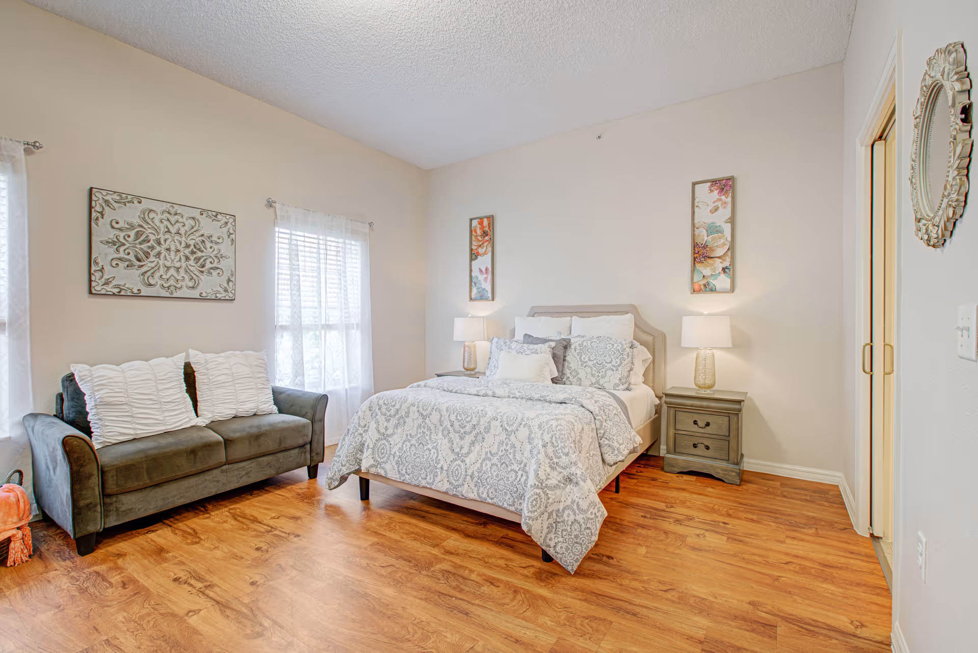 Sunny bedroom featuring a bed with patterned bedding, two nightstands and lamps, a loveseat, and wall art.