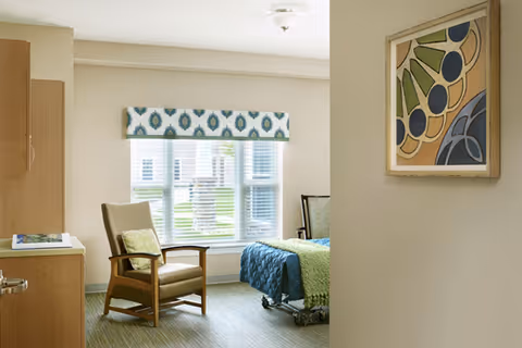 A bright and cozy bedroom in a senior living facility with a large window covered by a patterned valance. The room features a comfortable armchair with a cushion, a bed with blue and green bedding, a wooden cabinet, and a framed abstract artwork on the wall.